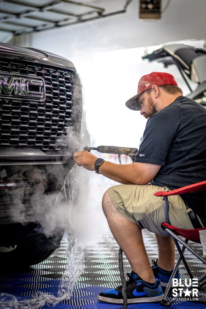 STEPH 8 9132024 | Blue Star Auto Salon Mechanic in a red cap using a heat tool on a car's front grille, steam rising in a workshop.
