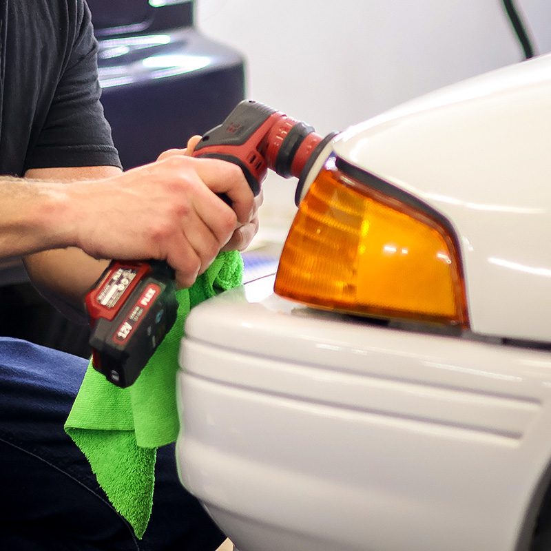 Person using a red cordless drill on the front bumper of a white car, with an orange headlight nearby and a green towel nearby.