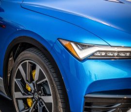 Blue sports car front-left corner with a large alloy wheel and yellow brake caliper, glossy blue paint, showroom lighting.