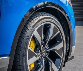Front-right wheel area of a blue car showing a large, multi-spoke alloy rim, yellow brake caliper, and a MichelIn tire with a glossy black fender trim.