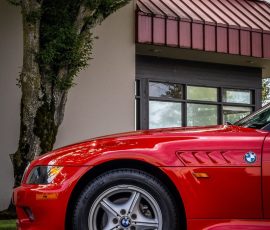 Red BMW sports car parked by a beige building with a large window, tree on the left, Blue Star Autosalon watermark.