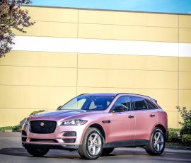 Pink Jaguar SUV parked in a lot in front of a pale yellow wall, with a tree on the left and BLUE STAR AUTOSALON logo in the corner.