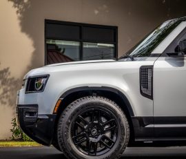 White SUV with large black multi-spoke wheels parked on dark pavement beside a beige building; BLUESTAR AUTOSALON logo in the bottom right corner.