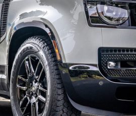Close-up of the front left section of a silver SUV, showing the headlight, grille, and large black alloy wheel with a Goodyear tire.