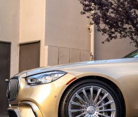 Front quarter view of a gold luxury car with large chrome multi-spoke wheels parked outdoors; Blue Star Autosalon watermark in lower right corner