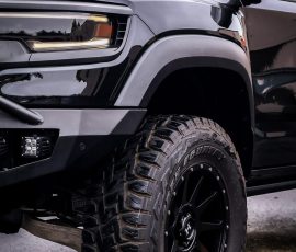 Close-up of a black pickup truck's front left corner with a large off-road tire and rugged fender, watermark visible.
