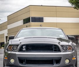 Front view of a gray Ford Mustang Shelby GT500 with a bold grille, parked in a lot near a beige building.
