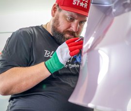 Bearded auto technician in a red BLUE STAR cap and gloves inspecting a white car panel in a bright workshop.