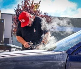 Man wearing a red cap and black shirt grinds a car hood, smoke rising around him, in an auto shop setting with a blue car. The 'BLUE STAR AUTOSALON' logo is visible on the car door.
