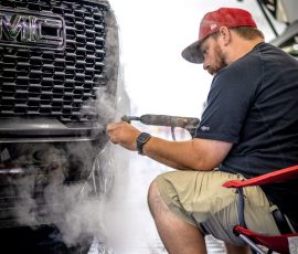Mechanic in a red cap using a heat tool on a car's front grille, steam rising in a workshop.