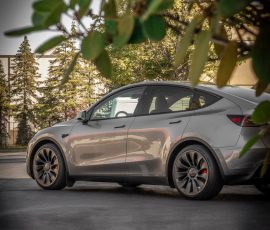 Gray electric SUV parked in a lot, photographed from the side with leafy foreground framing the scene; red brake calipers subtly visible; Blue Star Autosalon watermark in corner.
