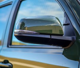 Close-up of a green car side mirror reflecting a building and blue sky, with the Blue Star Autosalon logo in the bottom-right corner.